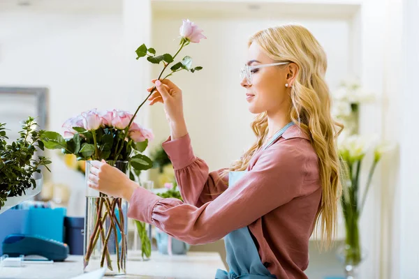 Beautiful female florist putting pink rose in bouquet in flower shop — Stock Photo