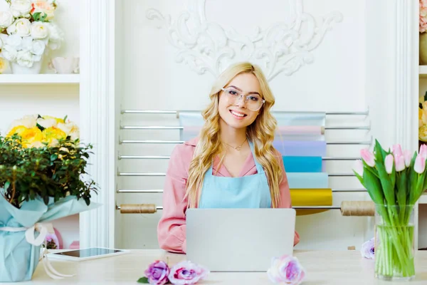Bella donna proprietario negozio di fiori in occhiali seduto alla scrivania, guardando la fotocamera e utilizzando il computer portatile — Foto stock