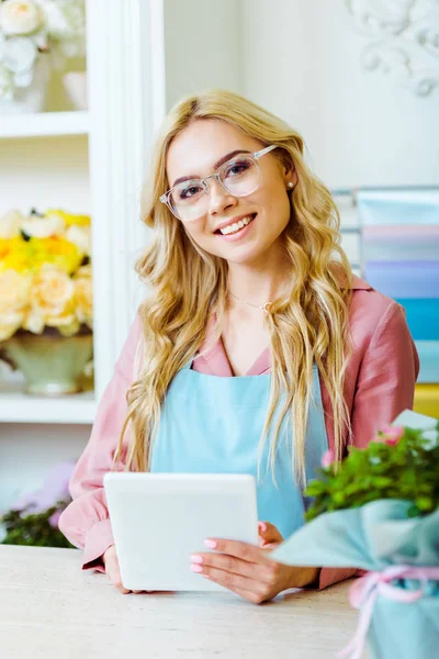 Schöne lächelnde Blumenverkäuferin mit Brille, die in die Kamera schaut und am Tresen ein digitales Tablet benutzt — Stockfoto