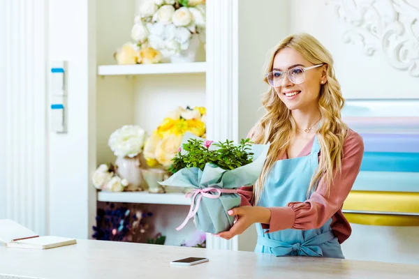 Hermosa florista femenina sonriente en gafas que presentan ramo en la tienda de flores - foto de stock