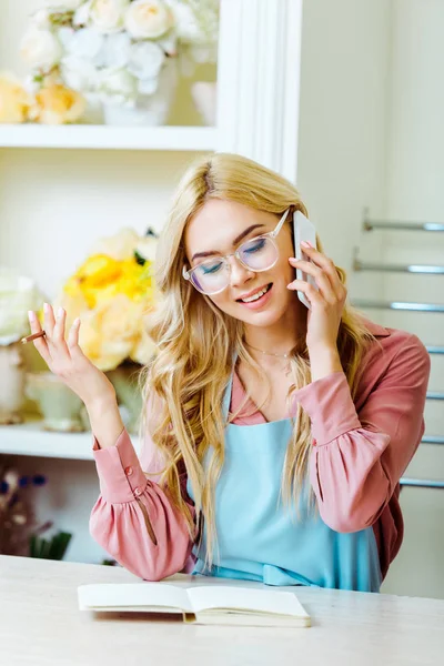 Beautiful smiling female flower shop owner in glasses talking on smartphone and holding pencil — Stock Photo