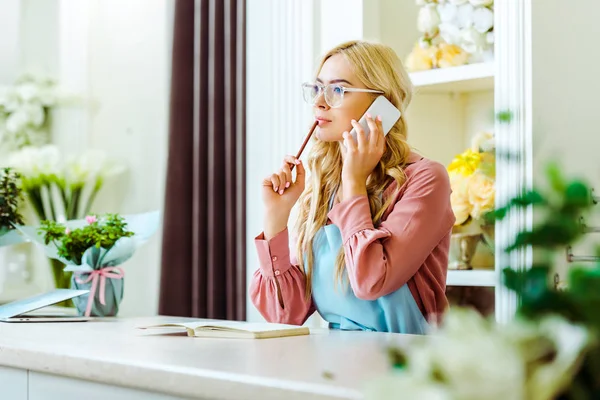 Beautiful female flower shop owner in glasses talking on smartphone and holding pencil — Stock Photo