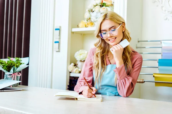 Bela sorridente dono da loja de flores do sexo feminino em óculos escrevendo em notebook e segurando smartphone — Fotografia de Stock