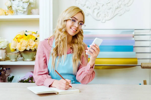 Красивая улыбающаяся владелица цветочного магазина в очках, пишущая в блокноте и используя смартфон — стоковое фото