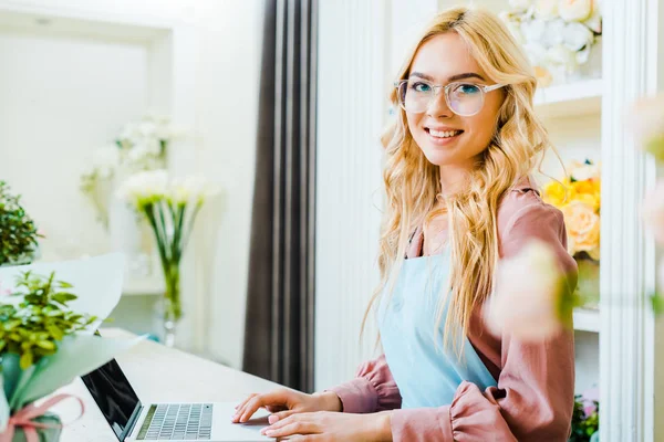 Beautiful female florist in glasses using laptop in flower shop and looking at camera — Stock Photo