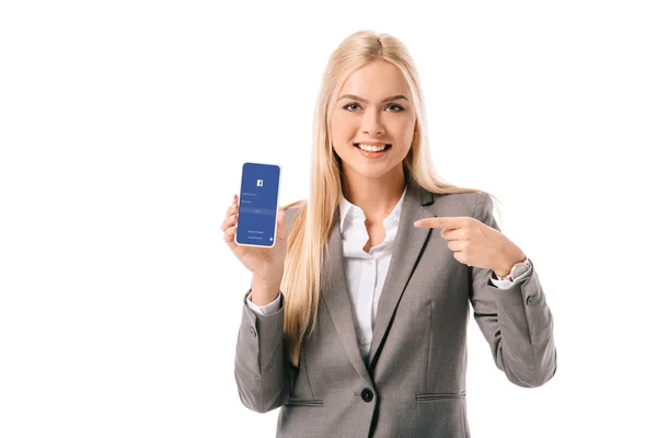 Улыбающаяся деловая женщина, указывающая на смартфон с помощью приложения facebook, изолированного на белом — стоковое фото