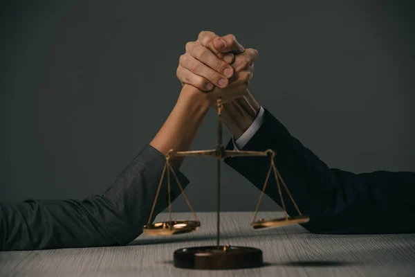 Partial view of businessman and businesswoman arm wretsling on wooden table with scales of justice on grey — Stock Photo