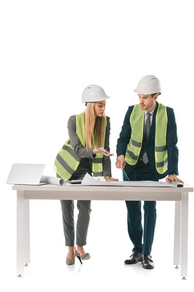 Arquitectos en chalecos de seguridad y cascos que trabajan con planos y portátil, aislados en blanco - foto de stock