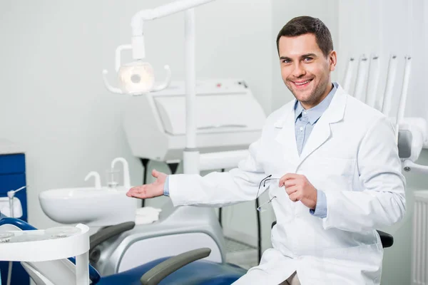 Guapo dentista sonriendo mientras gesticulando en la clínica - foto de stock