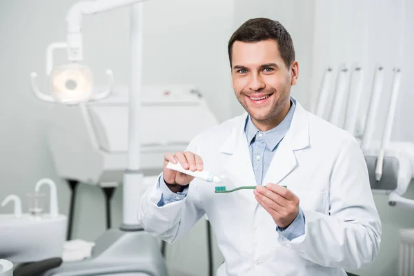 Улыбающийся дантист сжимает зубную пасту на зубной щетке в стоматологической клинике — стоковое фото