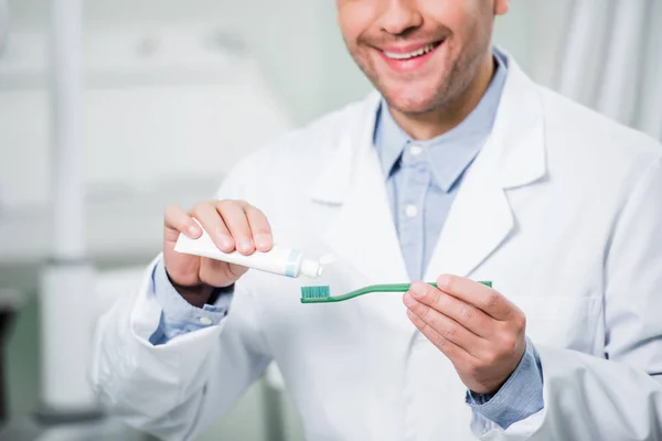 Vista recortada del dentista sonriente apretando la pasta de dientes en el cepillo de dientes en la clínica dental - foto de stock