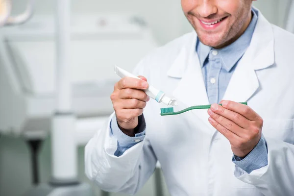 Vue recadrée du dentiste heureux serrant le dentifrice sur la brosse à dents dans la clinique dentaire — Photo de stock