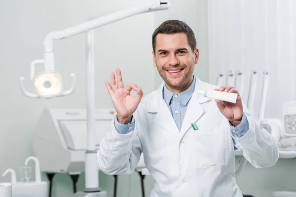 Zahnarzt hält Zahnpasta in der Hand und zeigt Okay-Zeichen in Zahnklinik — Stockfoto