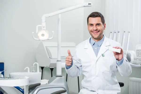 Dentista segurando pasta de dentes e mostrando polegar para cima na clínica odontológica — Fotografia de Stock