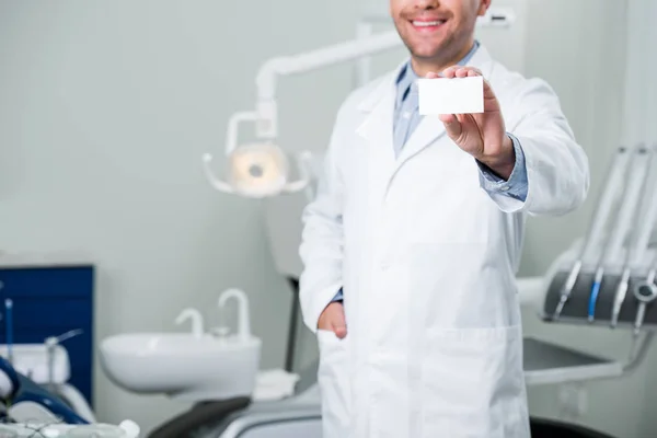 Vista recortada del dentista alegre de pie con la mano en el bolsillo y la celebración de la tarjeta en blanco - foto de stock