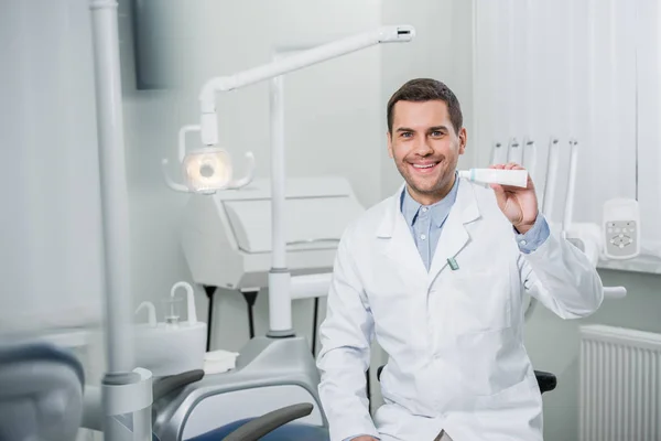 Dentista alegre segurando pasta de dentes sorrindo na clínica odontológica — Fotografia de Stock