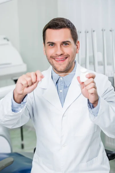 Alegre dentista de capa blanca sonriendo mientras sostiene hilo dental - foto de stock