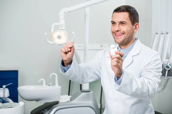 Schöner Zahnarzt im weißen Mantel lächelt, während er Zahnseide in der Hand hält — Stockfoto
