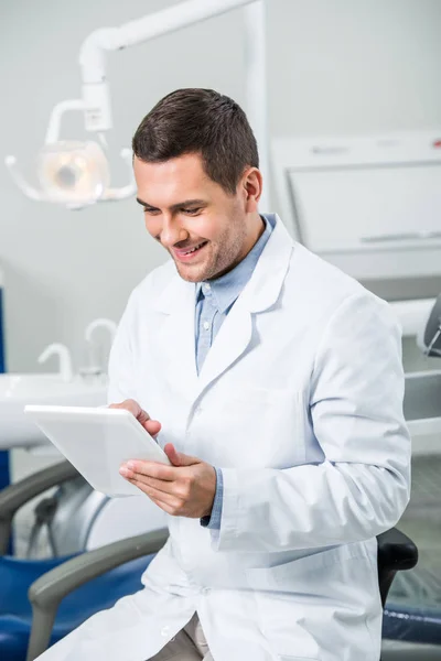 Dentista alegre em casaco branco segurando tablet digital na clínica odontológica — Fotografia de Stock