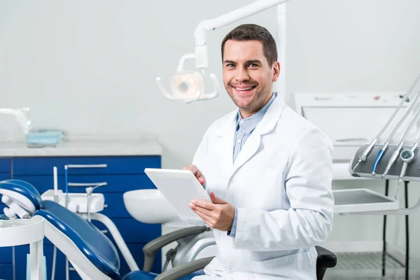 Dentista feliz de capa blanca sosteniendo tableta digital en la clínica dental - foto de stock