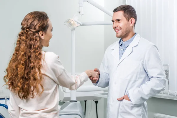 Dentista alegre de pie con la mano en el bolsillo y estrechando las manos con el paciente femenino - foto de stock