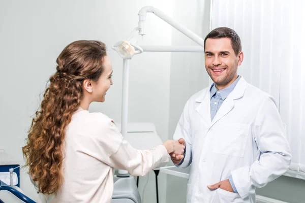 Lächelnder Zahnarzt steht mit der Hand in der Tasche und schüttelt Patientin die Hand — Stockfoto