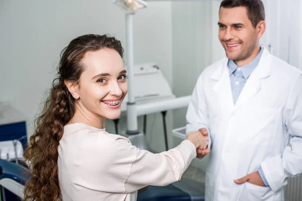 Messa a fuoco selettiva di donna allegra in bretelle stringendo la mano con dentista in piedi con mano in tasca — Foto stock