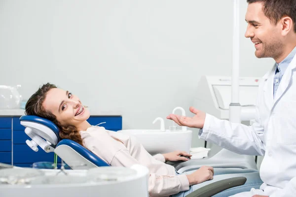 Donna allegra in apparecchio sorridente vicino bel dentista in clinica — Foto stock