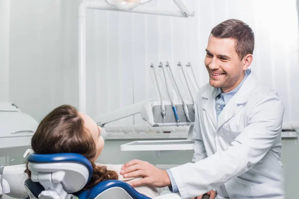 Apuesto dentista sonriendo mientras mira paciente - foto de stock