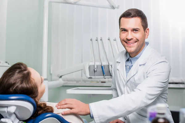 Guapo dentista en blanco abrigo sonriendo cerca de mujer paciente - foto de stock