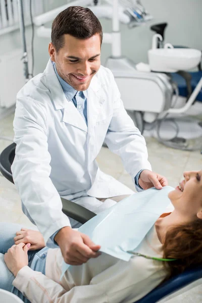 Beau dentiste en manteau blanc souriant près du patient attrayant — Photo de stock