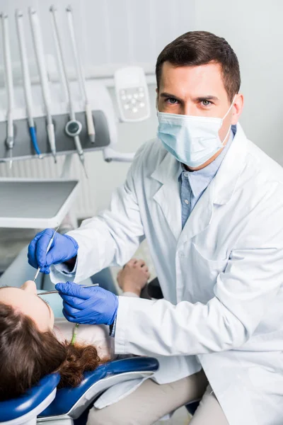 Dentista en guantes de látex y máscara que sostiene el equipo dental cerca del paciente femenino - foto de stock