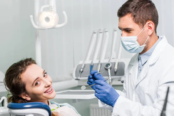 Dentista in lattice guanti e maschera in possesso di attrezzature dentali vicino bella donna con bretelle — Foto stock