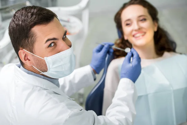 Selektiver Fokus des Zahnarztes in der Maske, die die zahnärztliche Ausrüstung während der Untersuchung der Patientin hält — Stockfoto