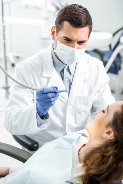 Messa a fuoco selettiva del dentista in mantello bianco e maschera che tiene lo strumento dentale vicino al paziente femminile — Foto stock