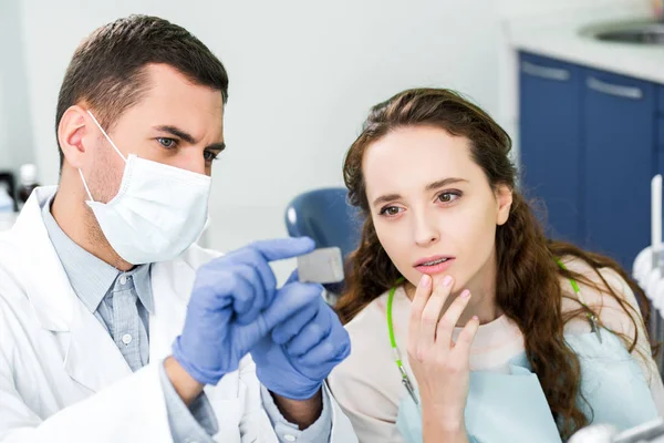 Enfoque selectivo de la mujer pensativa mirando la radiografía de los dientes en las manos del dentista en guantes de látex y máscara - foto de stock