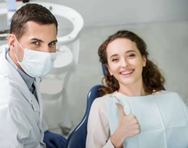 Messa a fuoco selettiva del dentista in maschera vicino sorridente paziente mostrando pollice verso l'alto — Foto stock