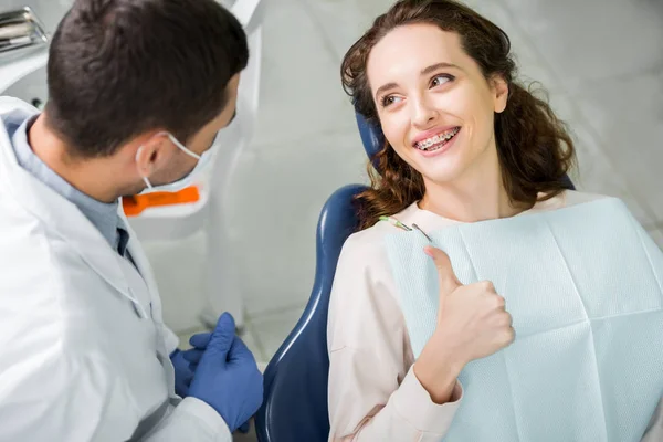Foyer sélectif de la femme dans les appareils souriants tout en montrant le pouce près du dentiste lors de l'examen — Photo de stock