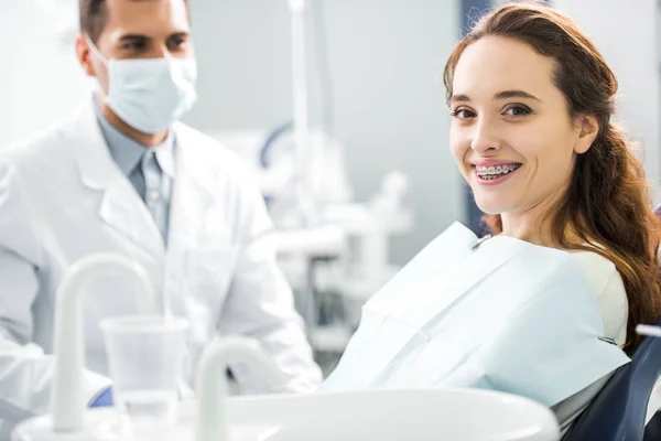 Enfoque selectivo de la mujer en frenillos sonriendo con el dentista de pie en la máscara en el fondo - foto de stock