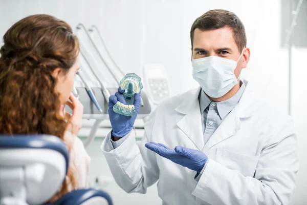 Selektiver Fokus des Zahnarztes in Maske und Latexhandschuhen, die das Zahnmodell in der Nähe der Patientin zeigen — Stockfoto