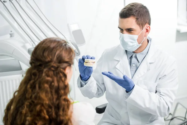Селективный фокус стоматолога в маске и латексных перчатках, показывающих модель зубов рядом с женщиной — стоковое фото
