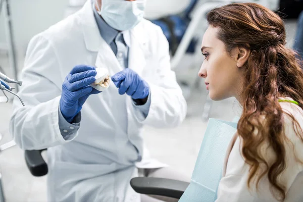 Vista recortada del dentista en guantes de látex sosteniendo los dientes modelo cerca del paciente femenino - foto de stock