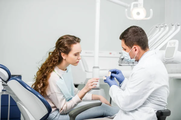 Dentist in latex gloves and mask holding teeth model near woman — Stock Photo