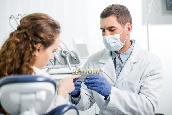 Donna che punta con il dito alla tavolozza dei colori dei denti vicino al dentista in maschera e guanti in lattice — Foto stock