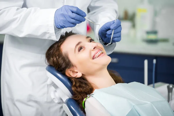 Обрізаний вид на стоматолога в латексних рукавичках, який вивчає веселу жінку в брекетах — стокове фото