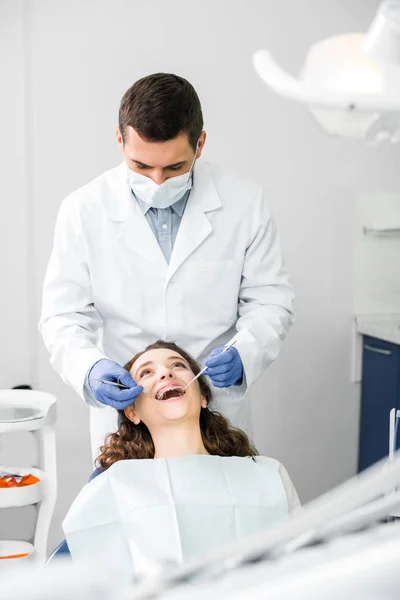 Dentiste en gants de latex examinant femme attirante dans des bretelles avec bouche ouverte — Photo de stock