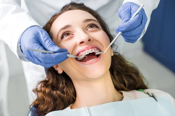 Vue recadrée du dentiste en gants de latex examinant femme gaie dans des bretelles avec bouche ouverte — Photo de stock