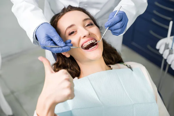 Vista recortada del dentista en guantes de látex examinando a una mujer atractiva con frenos con la boca abierta mostrando el pulgar hacia arriba - foto de stock