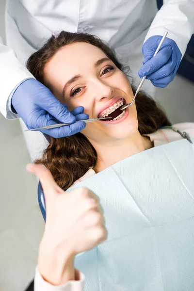 Vista recortada del dentista en guantes de látex examinando mujer alegre en frenos con la boca abierta mostrando el pulgar hacia arriba - foto de stock