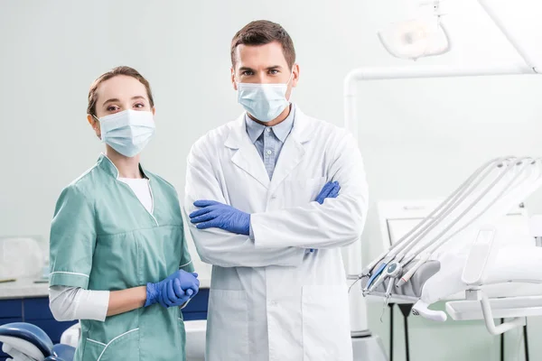 Dentistas em máscaras de pé com braços cruzados na clínica odontológica — Fotografia de Stock